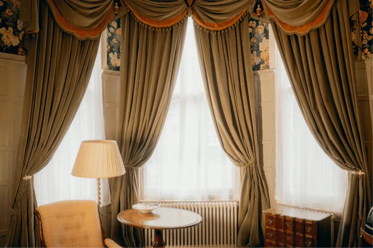 An elegant interior scene featuring a set of ornate drapes with a matching swaged valance, tied back from a large window. The curtains are a muted taupe-brown colour with a warm orange-gold trim along the top and made to measure by Sew Chic Interiors of Brighton & Hove