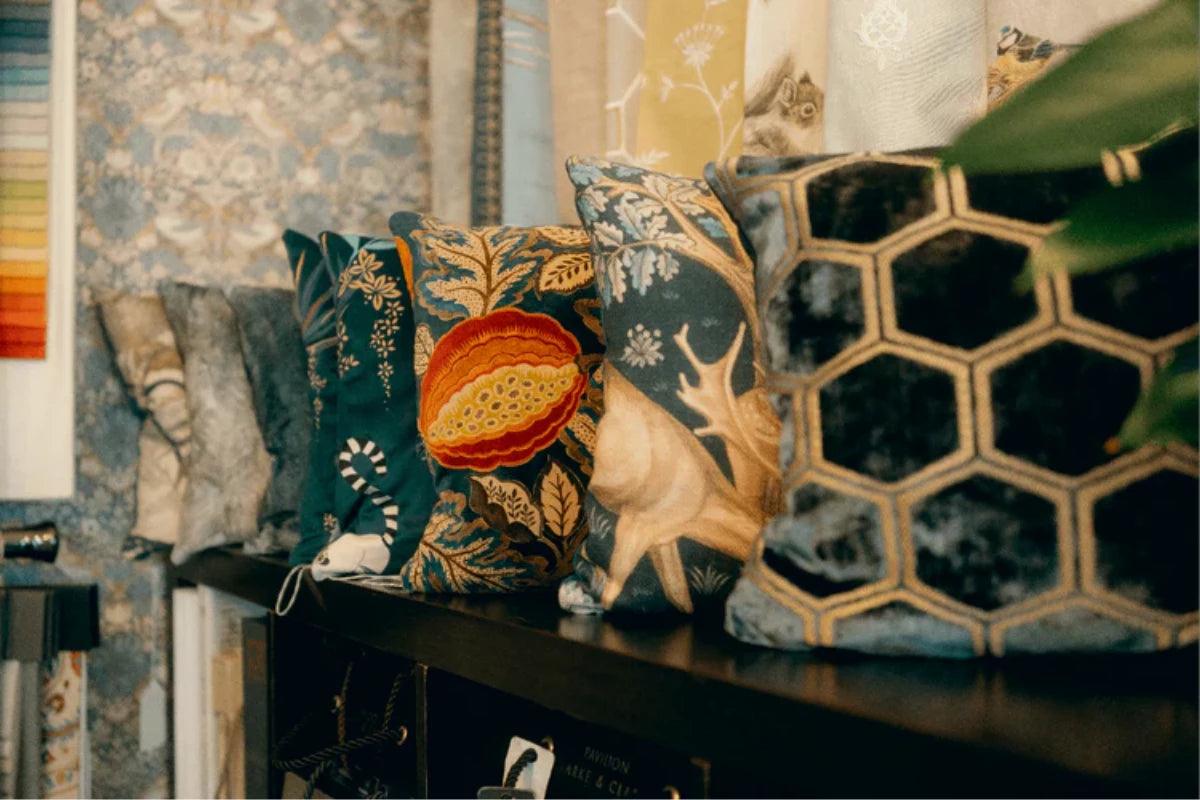 A collection of decorative throw pillows displayed on a dark wooden shelf in a styled room setting. The pillows feature various patterns including a prominent black and gold honeycomb design, a cream stag motif on a dark blue-green background, and a central orange pumpkin motif on a dark teal background all made by hand by Sew Chic Interiors of Brighton & Hove