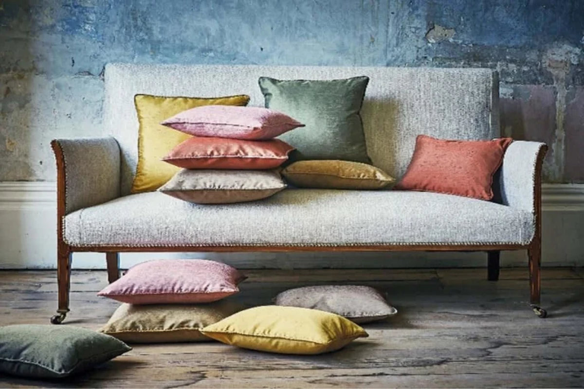 A light beige upholstered armchair with dark wood legs, styled with multiple decorative throw pillows in yellow, pink, green, and terracotta, set against a rustic blue-grey textured wall and wooden floor showing Soft Furnishings and Cushions made by Sew Chic Interiors of Brighton and Hove