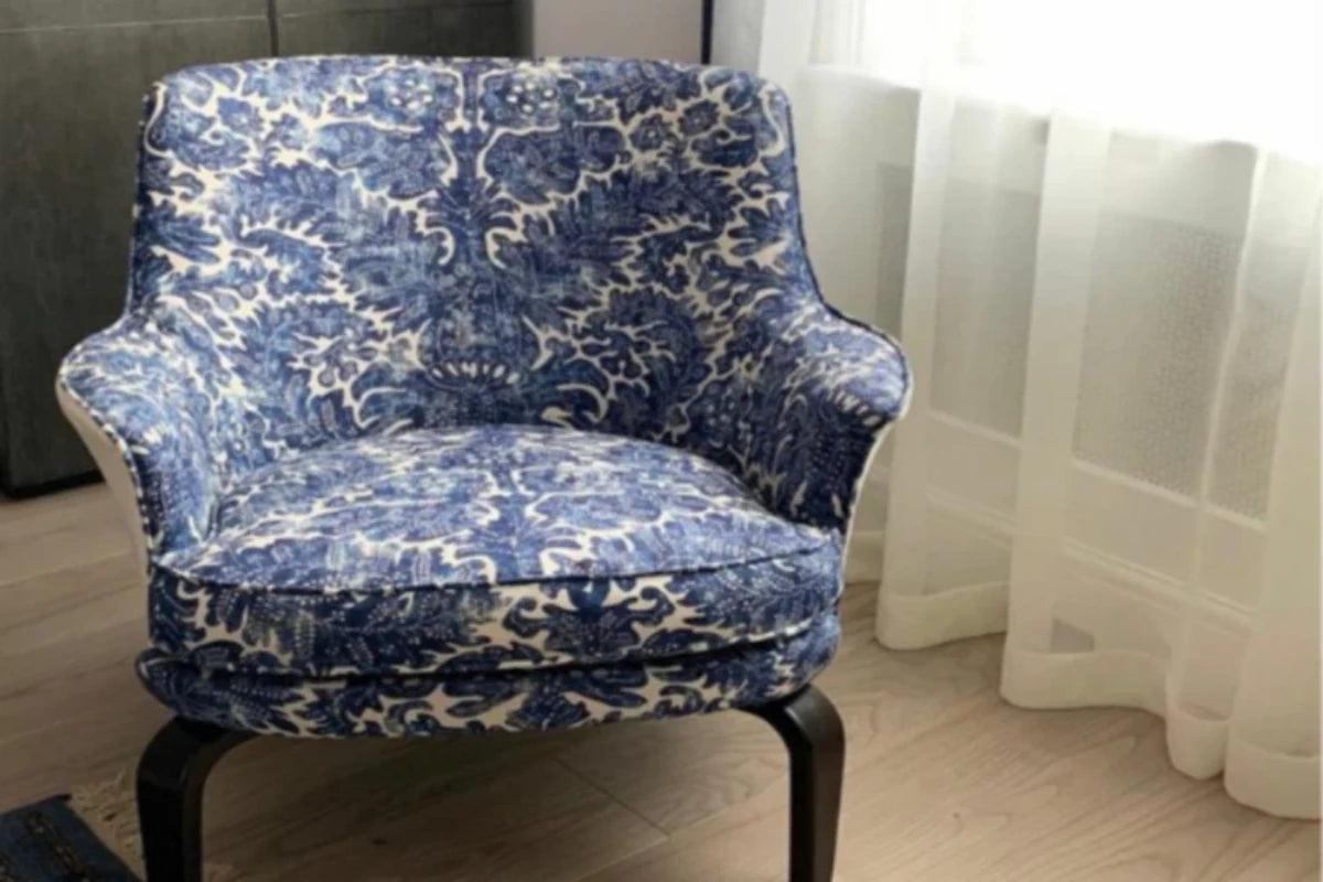 A single upholstered accent chair with a blue and white damask-style pattern, shown indoors near a window with sheer white curtains and light wood flooring re-upholstered by Sew Chic Interiors in Brighton & Hove
